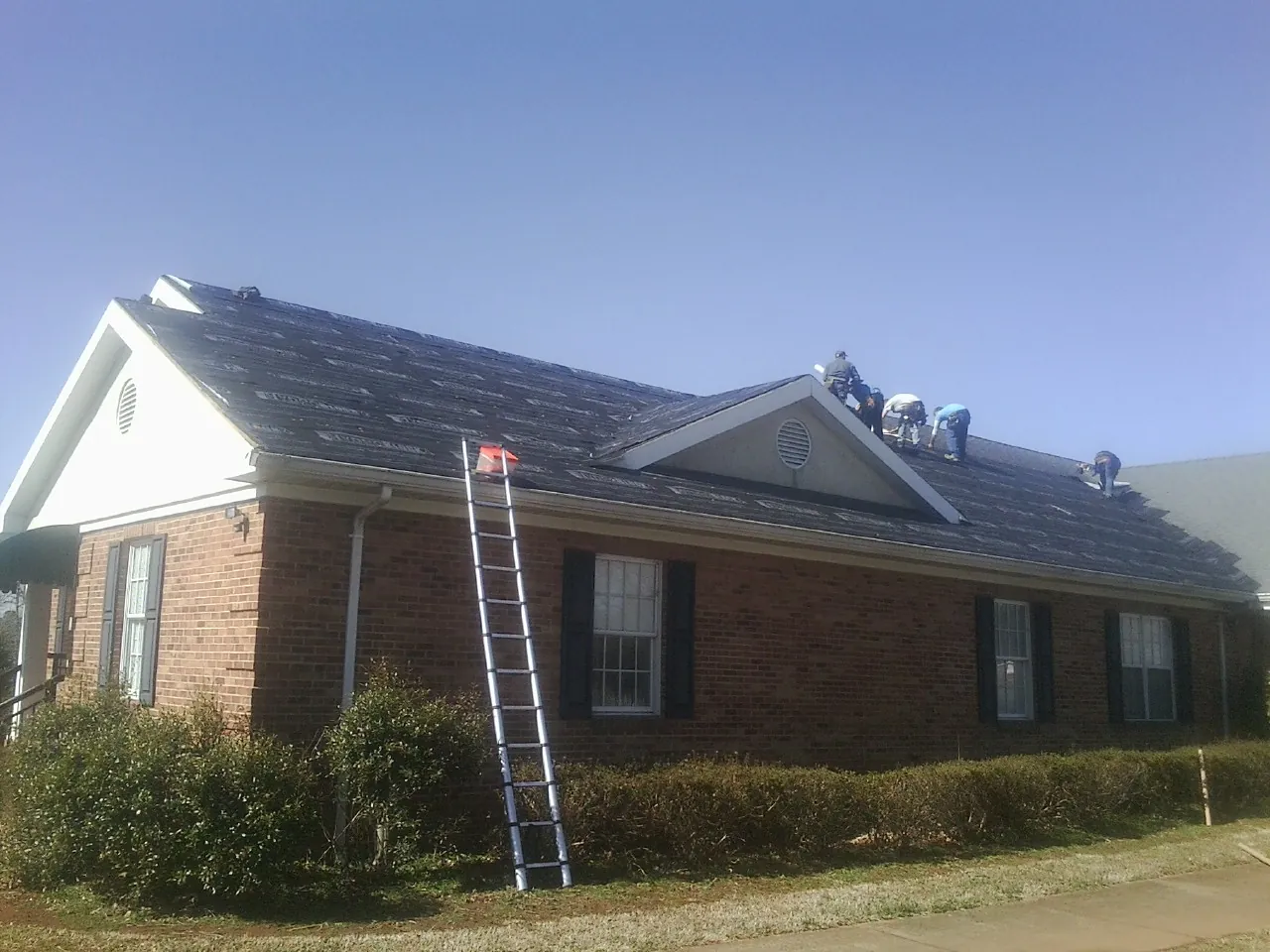 Crew tearing shingles off a brick ranch roof with a ladder set — Metal Roof Repair work by our crew in Bidwell