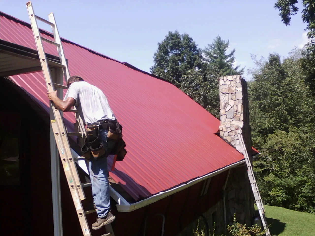 Roofer on a ladder beside a red metal roof and stone chimney — Soffit & Fascia Repair by Roofing Brothers in Bidwell
